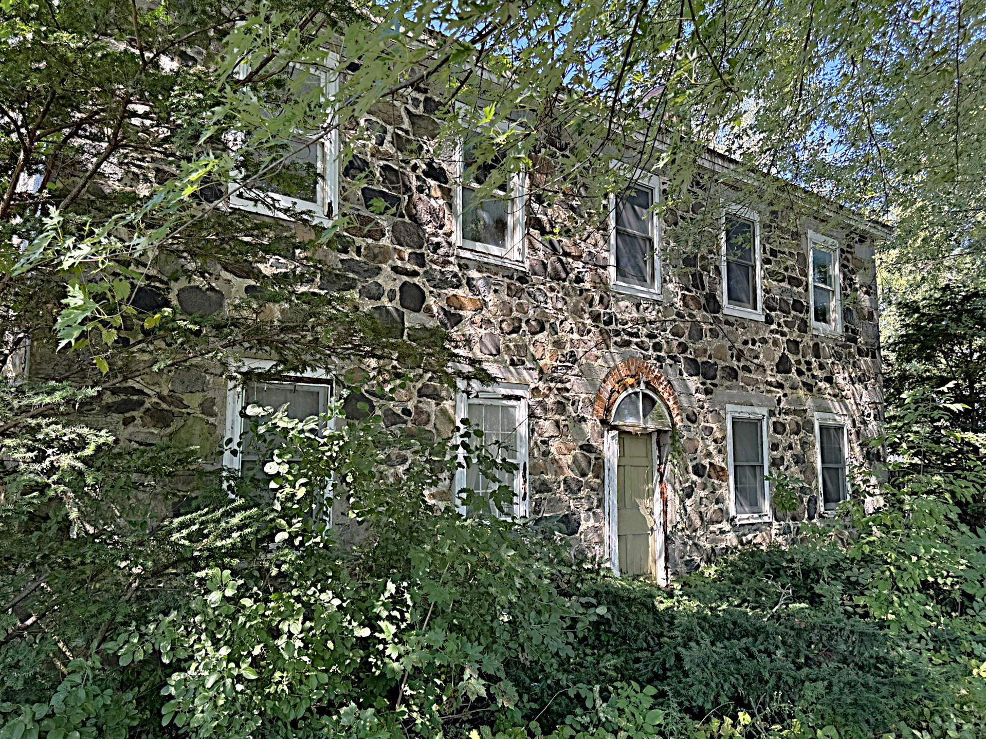 Two-story stone house nestled in dense greenery, with white-trimmed windows and an arched front entrance.