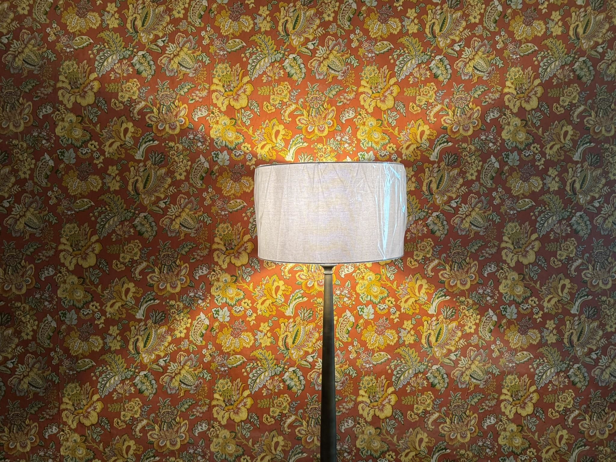Floor lamp with a white shade against vivid red floral wallpaper.