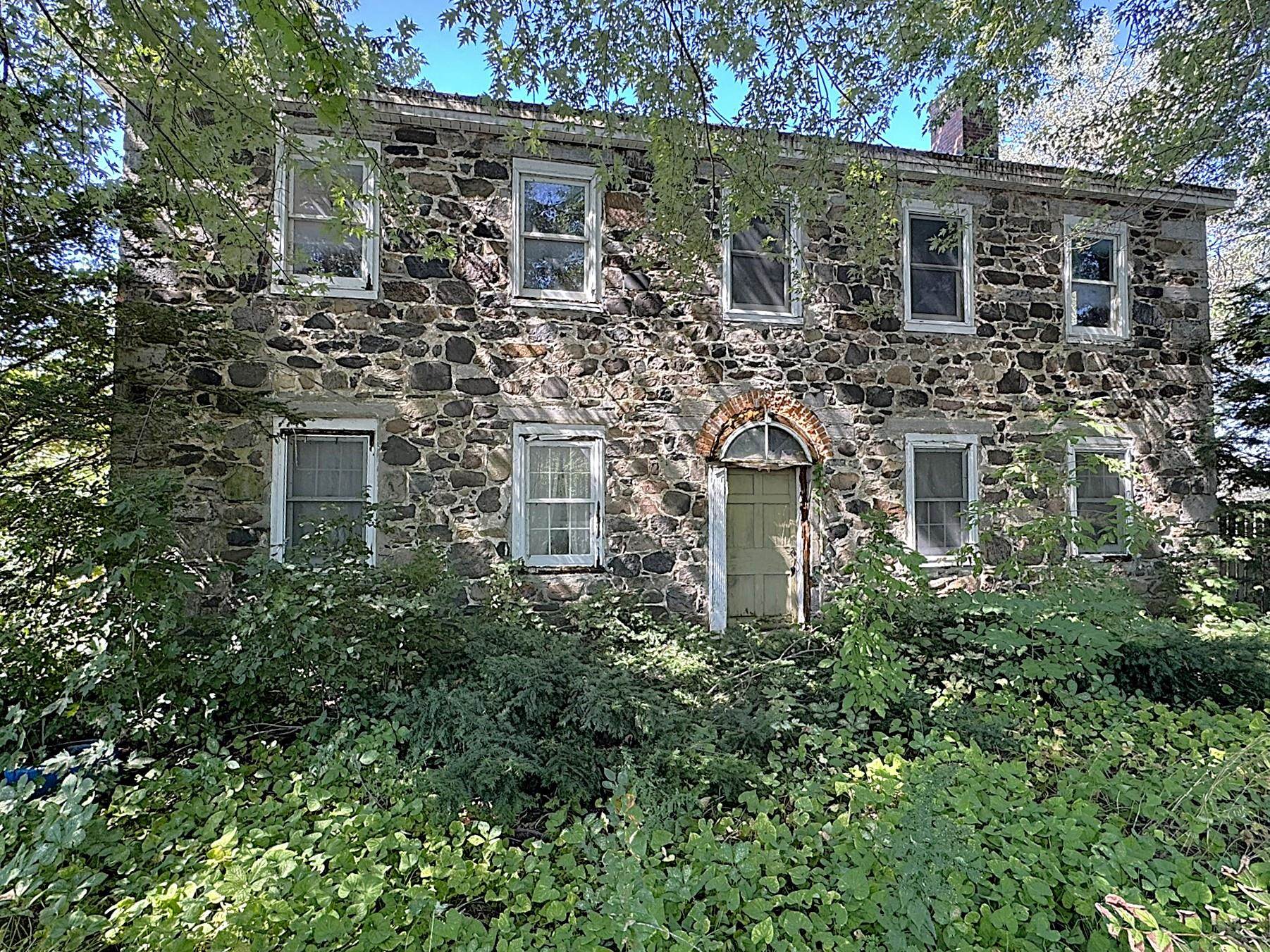 Two-story stone house with an arched brick entry, partly screened by dense greenery.