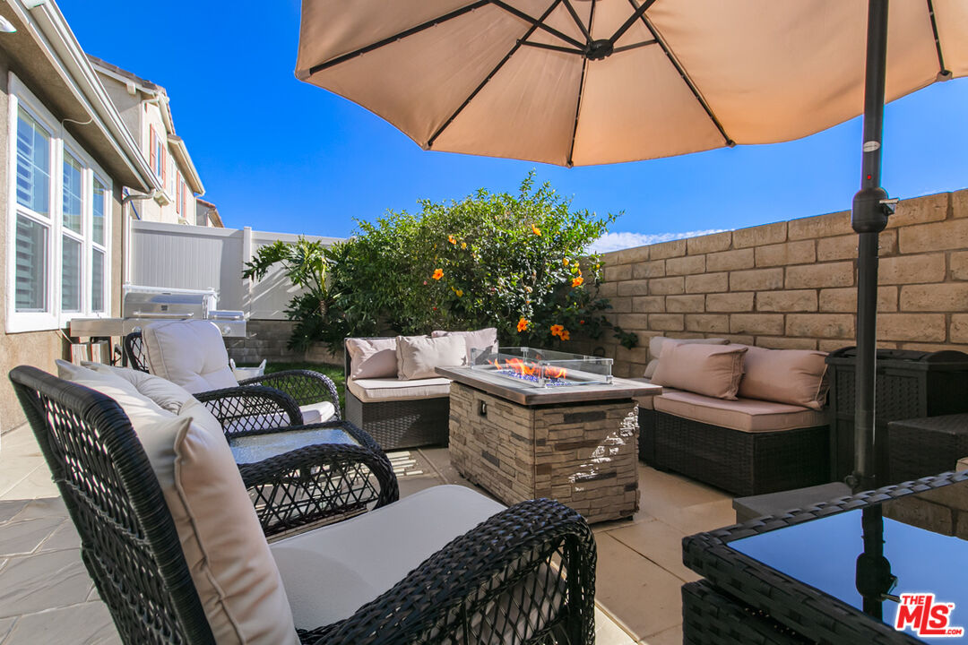 Patio with woven seating, a stone fire table, flowering shrub, and large umbrella.