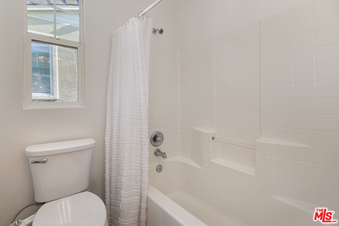 Bathroom with a white tub and tiled surround, ruffled shower curtain, toilet, and high window.