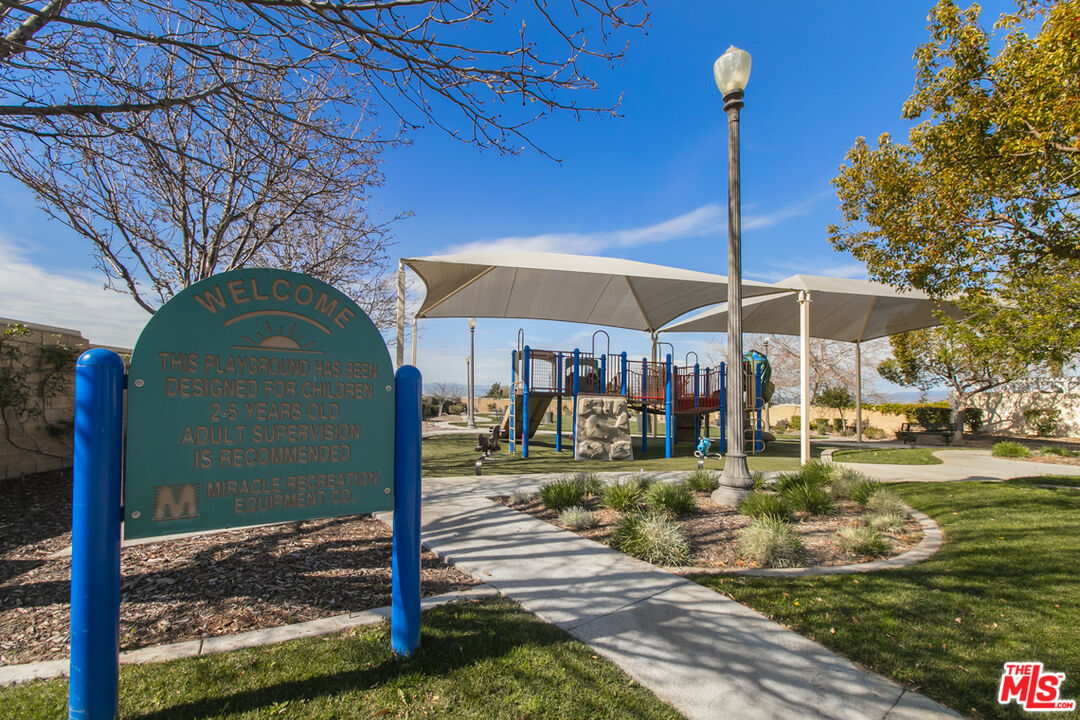 Community playground with a shaded climbing structure, paved path, and welcome sign.