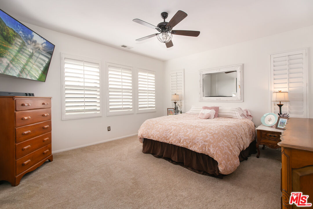 Bedroom with a pale pink floral bedspread, plantation shutters, and a dark ceiling fan.