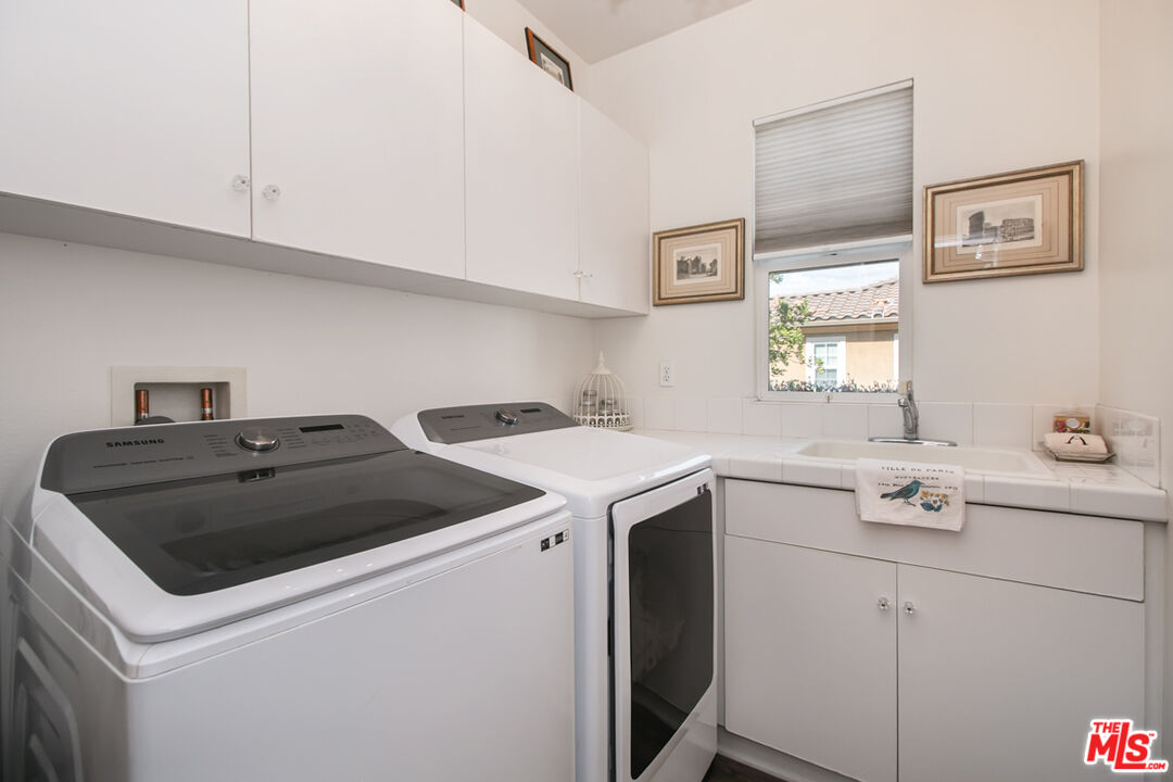 Laundry room with Samsung washer and dryer, white cabinetry, and a tiled utility sink beneath a window.