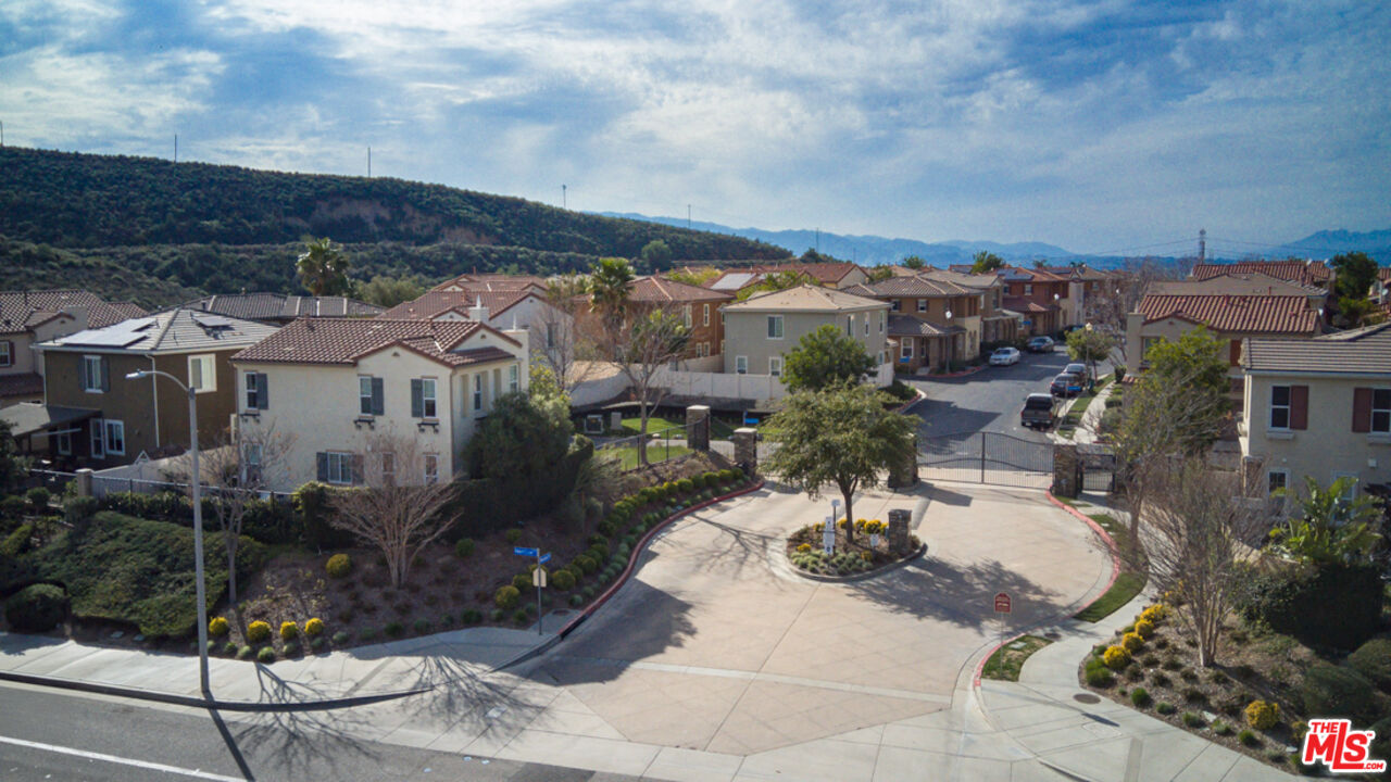 Aerial view of a gated neighborhood entrance and homes, with wooded hills behind.