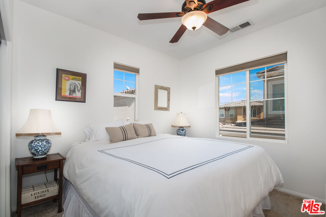 Furnished bedroom with a white bed, blue-trimmed bedding, and windows overlooking neighboring homes.
