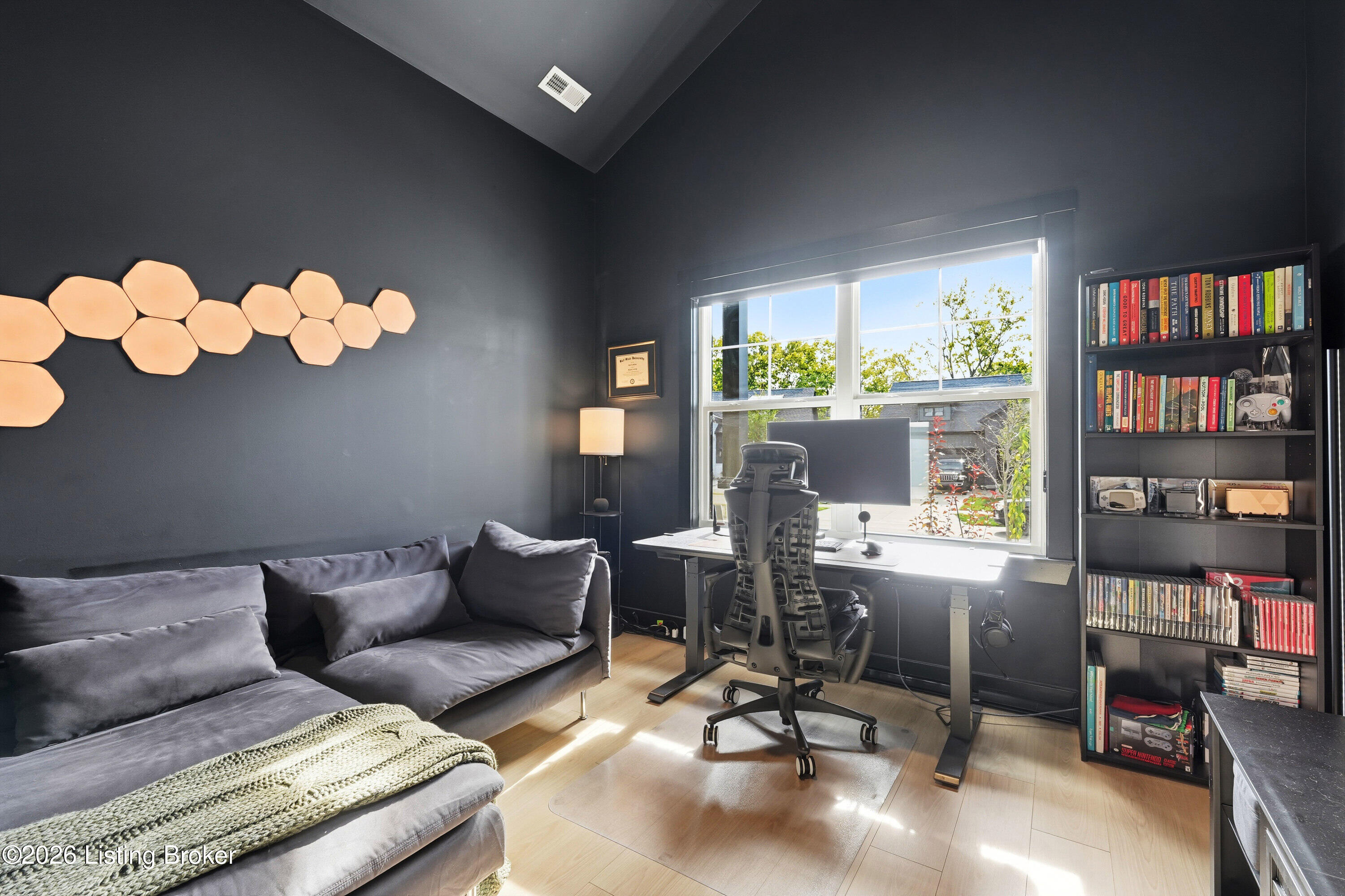 Home office with a gray sofa, desk by a large window, and bookshelves.