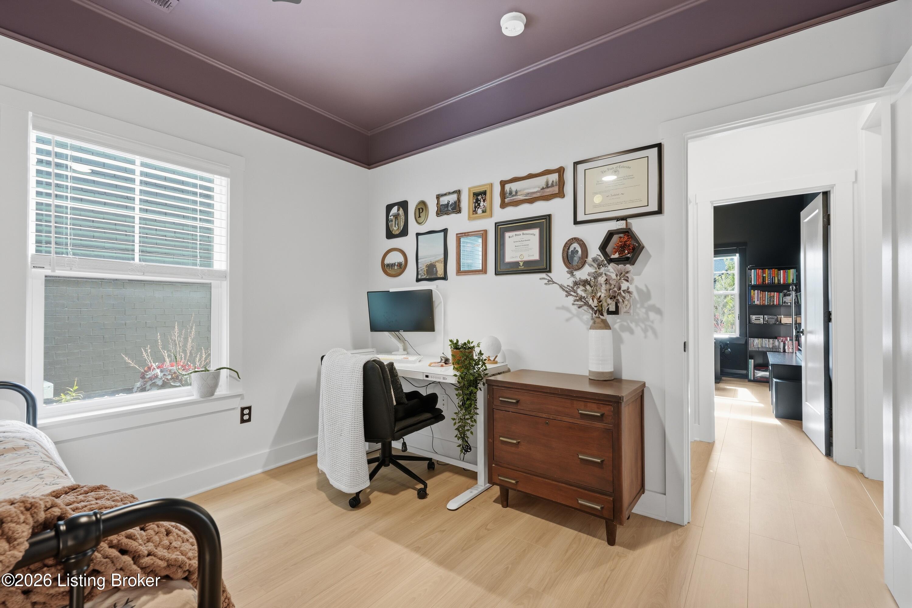 Bedroom with a compact desk workspace, gallery wall, and doorway to a dark book-lined room.