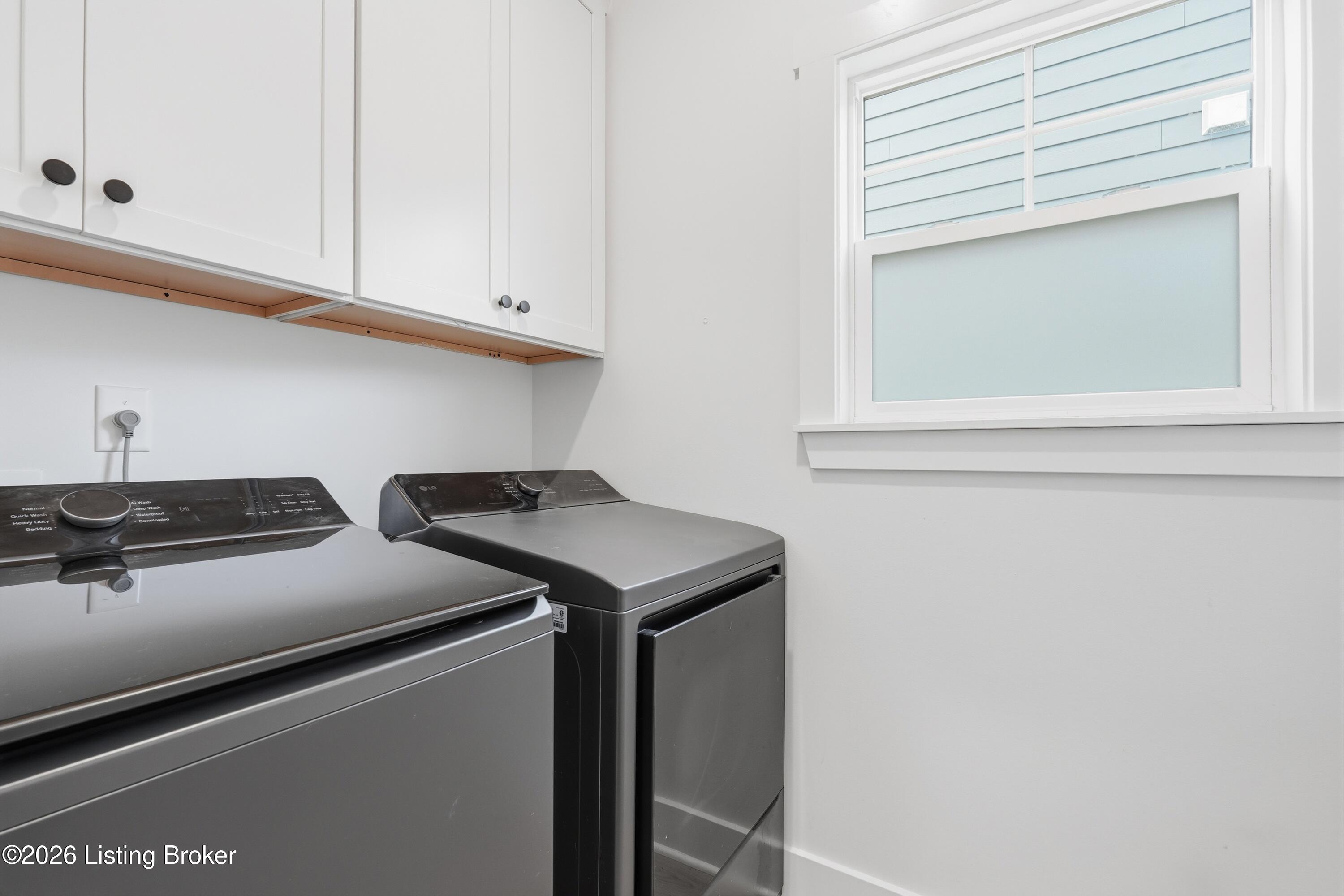 Laundry room with two dark appliances, white cabinets, and a window.