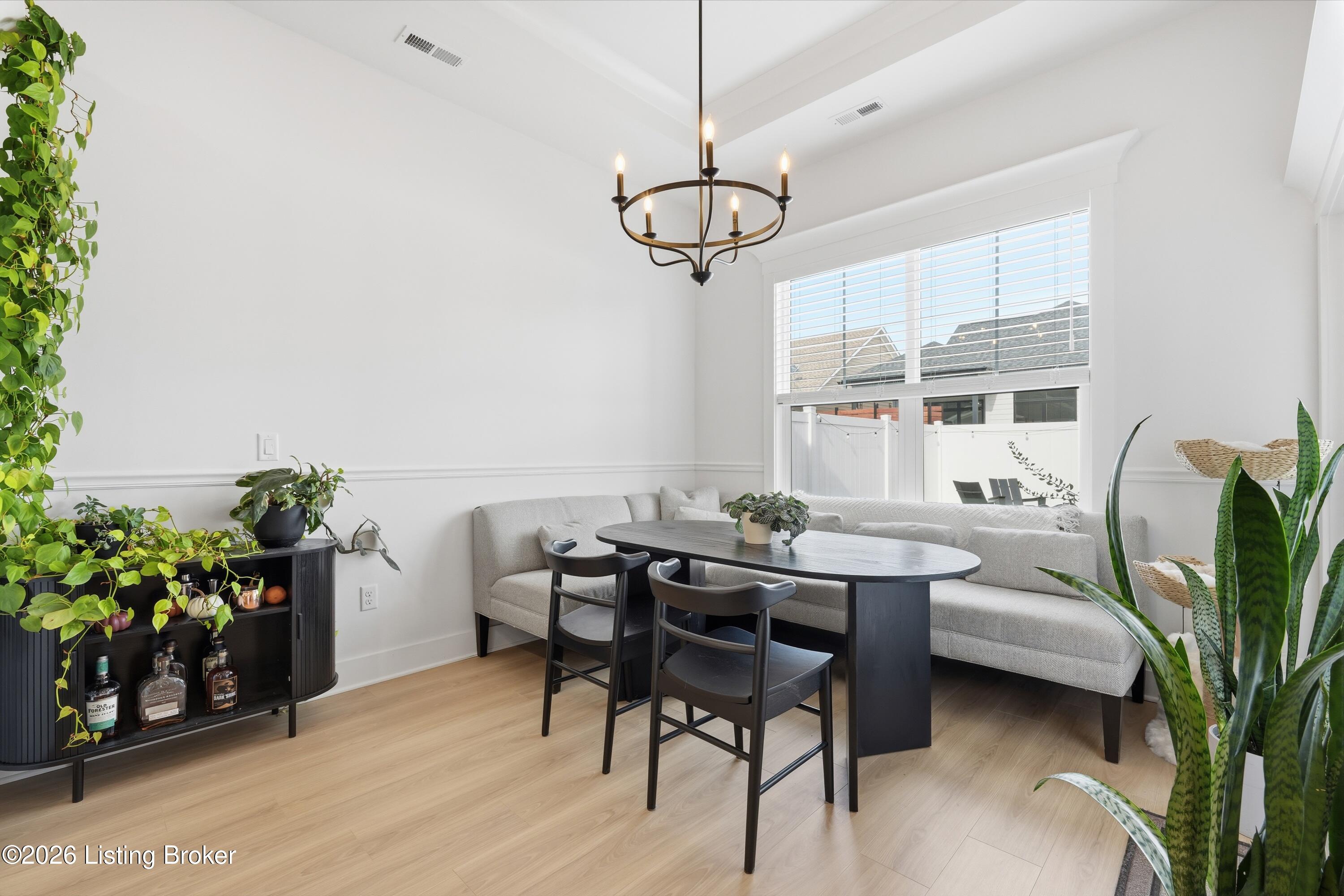 Dining area with a gray banquette, dark table, black chandelier, and leafy plants.