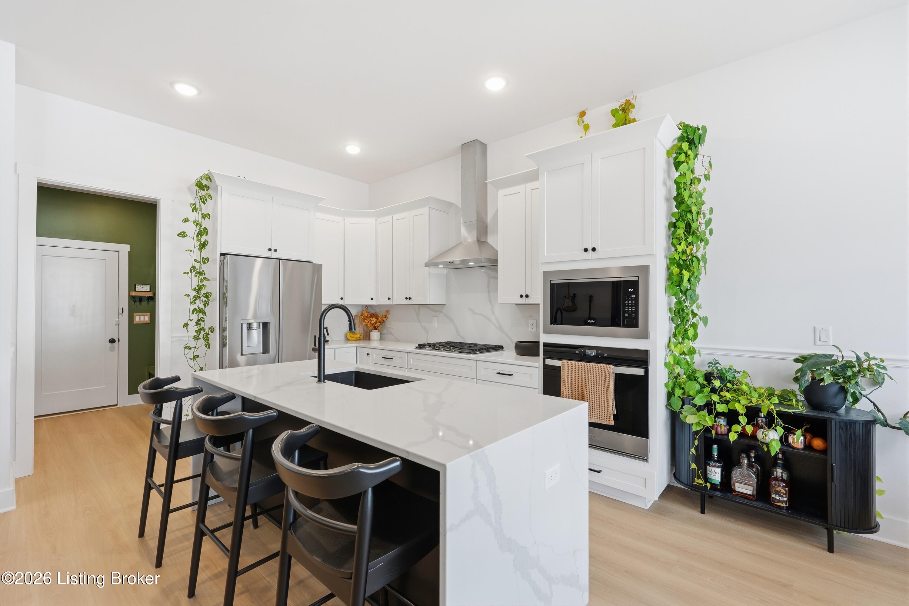 Bright white kitchen with a veined island, black stools, stainless appliances, and trailing plants.