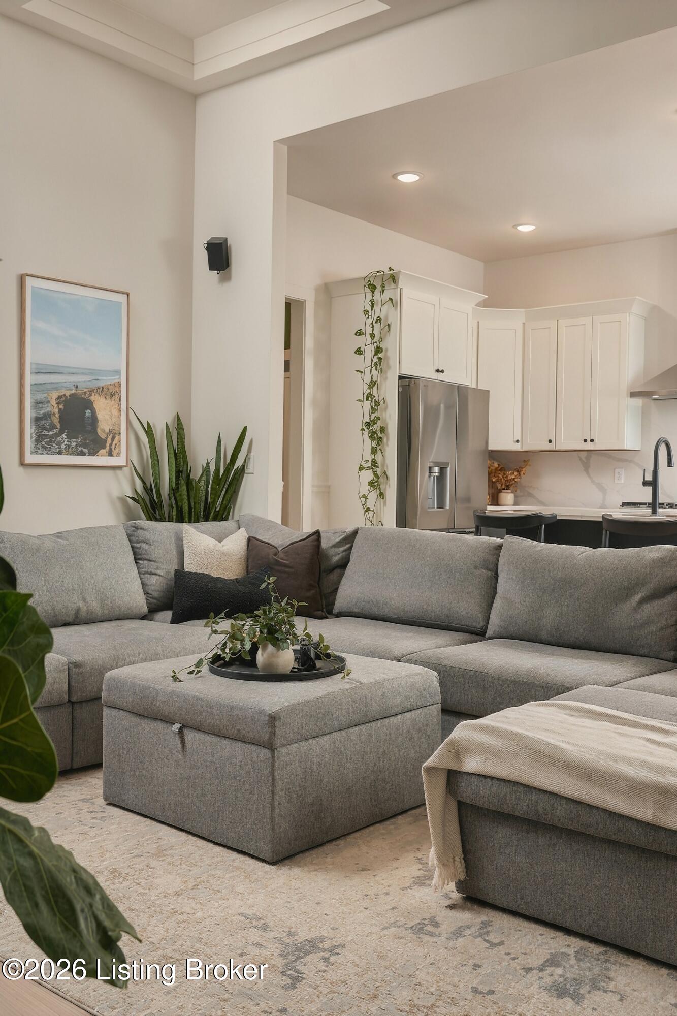 Living room with a gray sectional and ottoman opening into a white-cabinet kitchen.