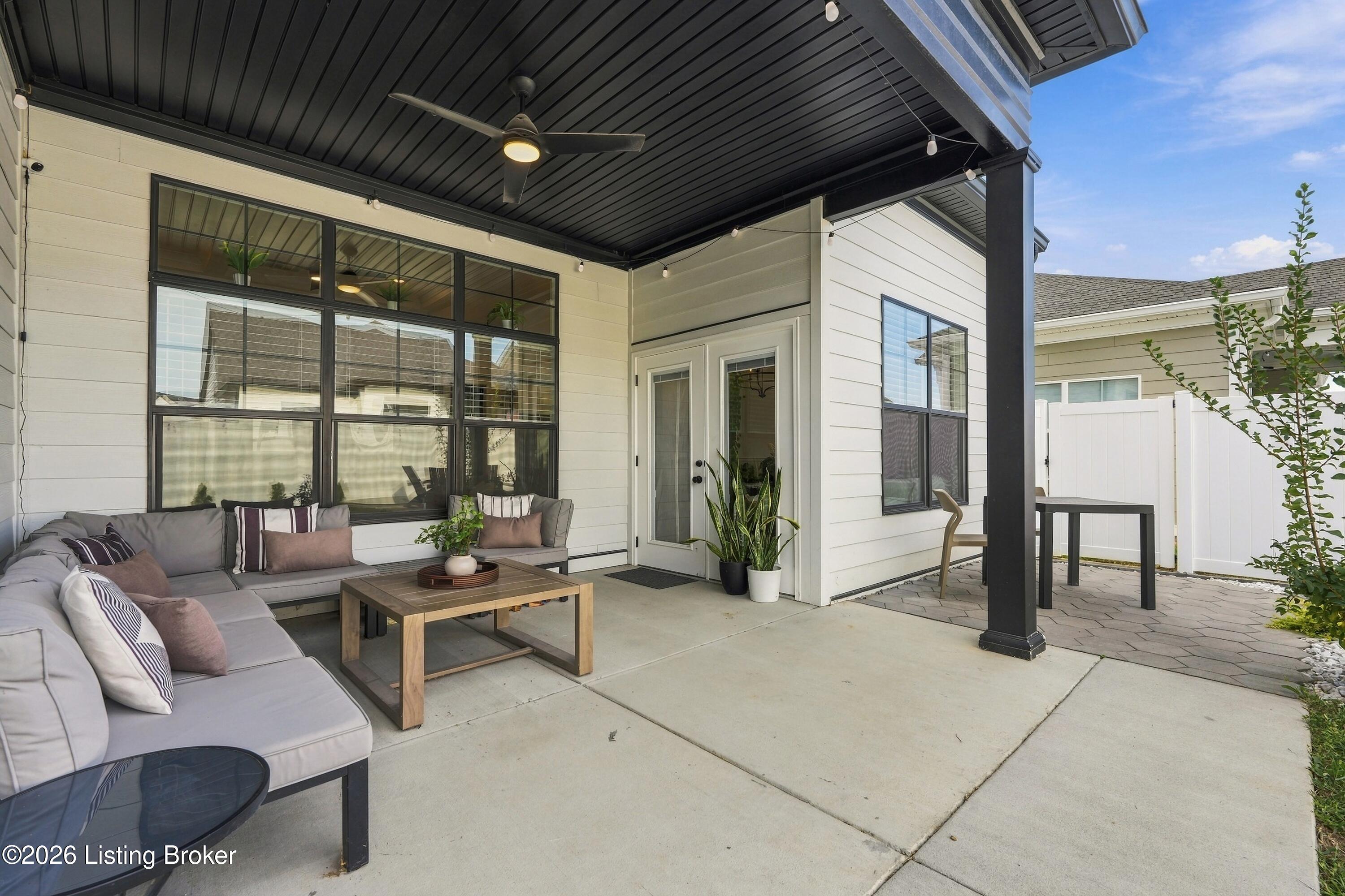 Covered patio with a gray sectional, coffee table, ceiling fan, and fenced paver seating area.