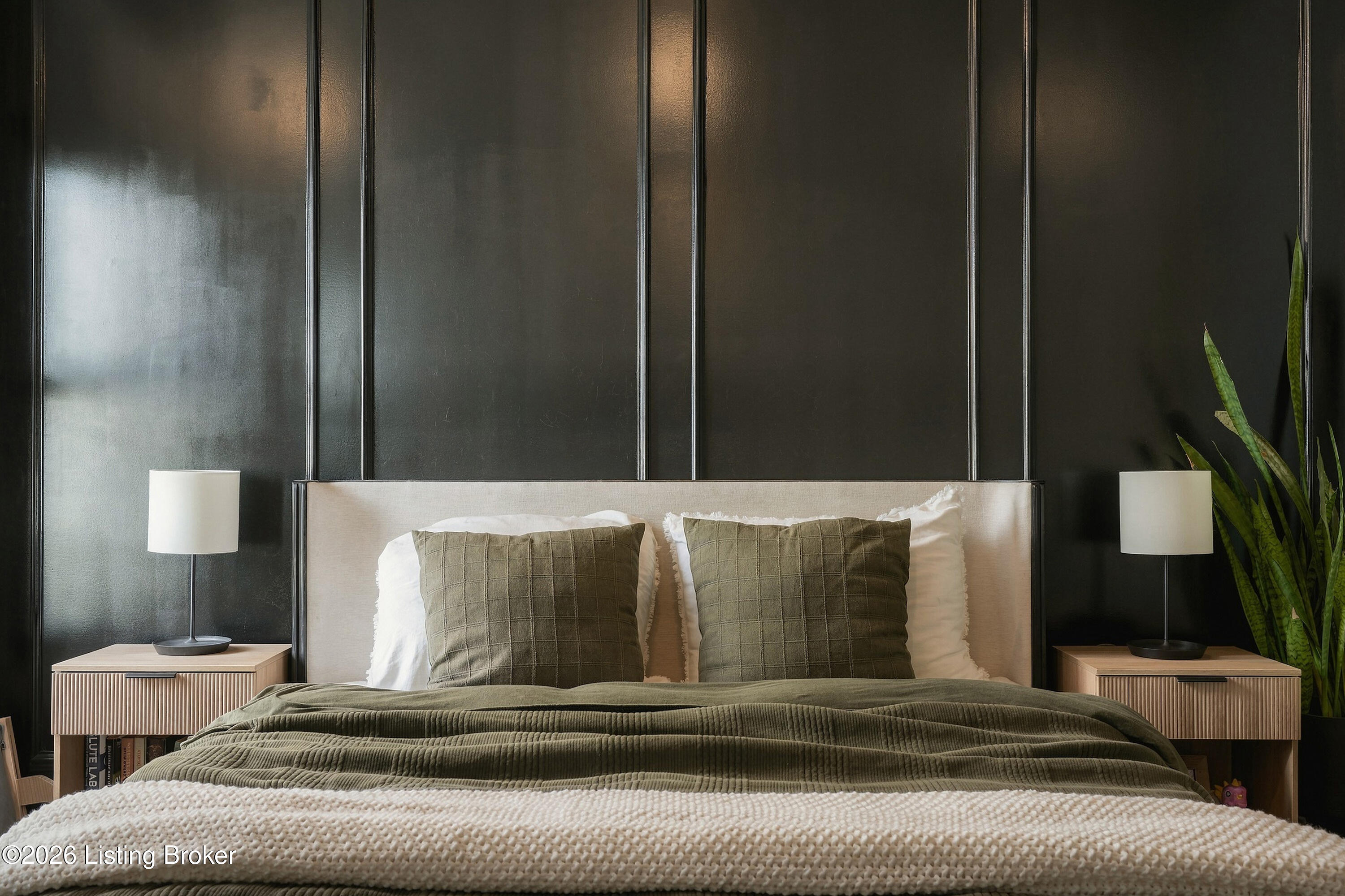 Bedroom with a large bed, olive bedding, dark paneled wall, matching bedside tables, and a tall snake plant.