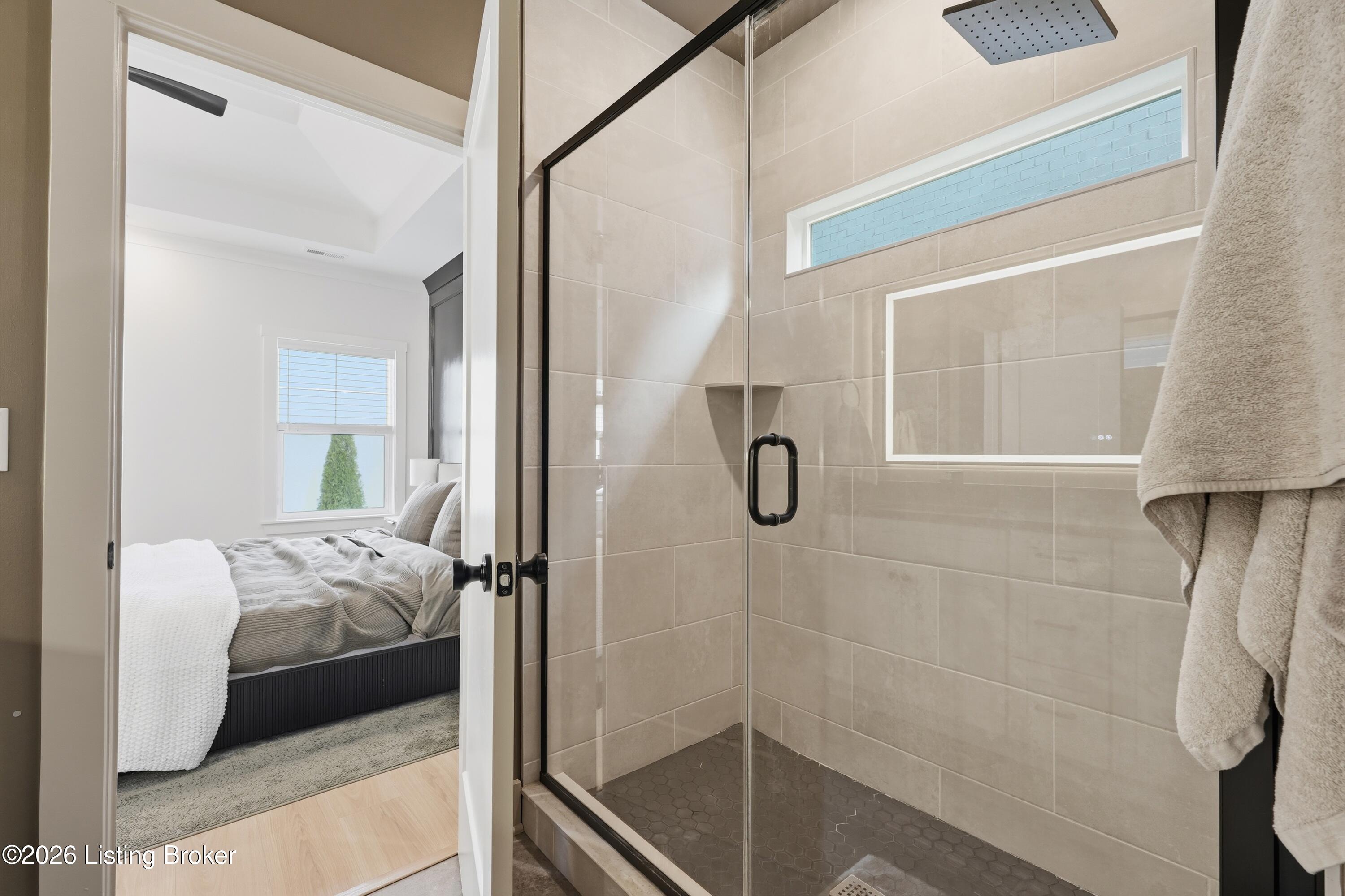 Walk-in tiled shower with black fixtures, viewed beside a doorway to a furnished bedroom.