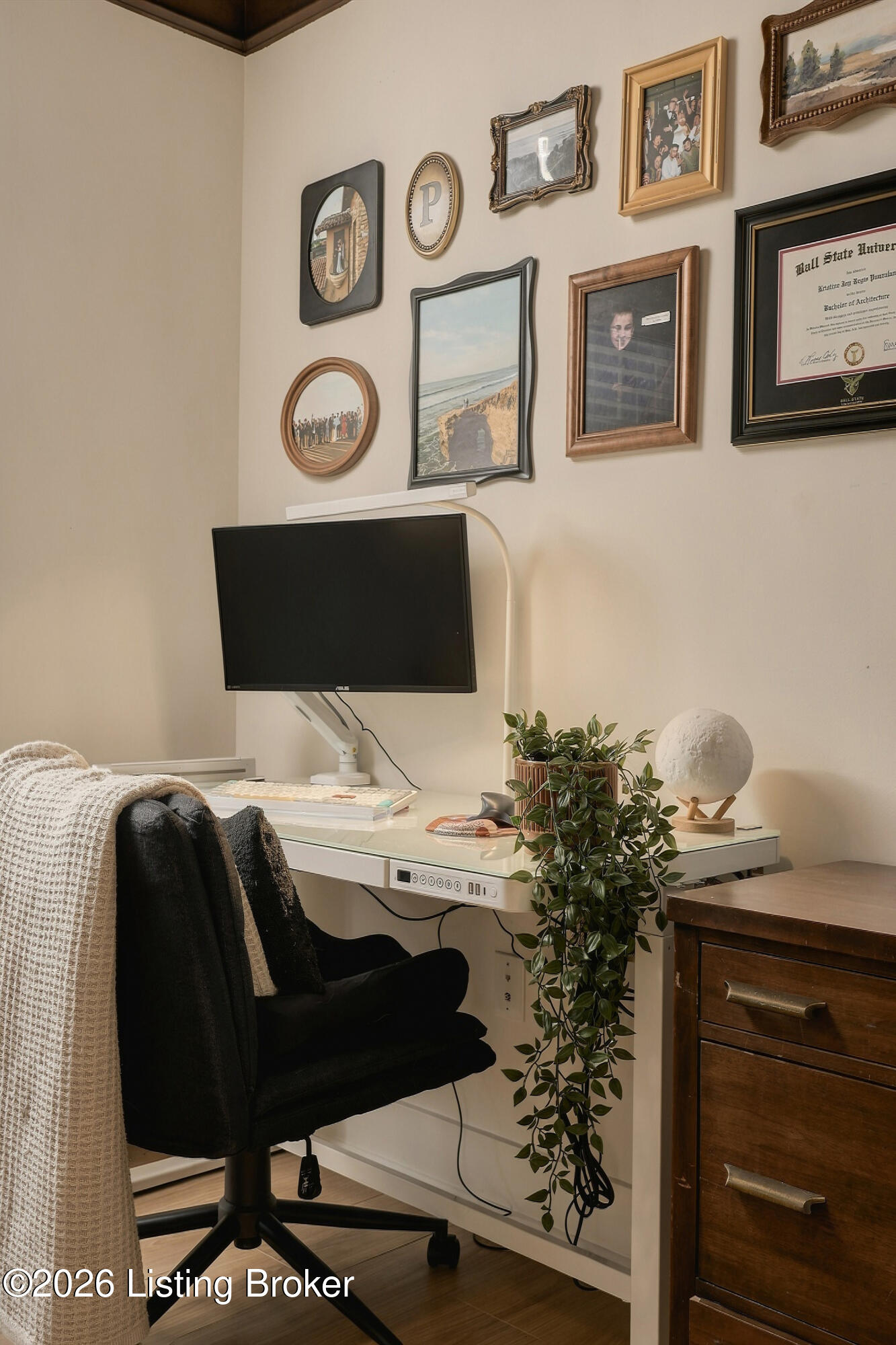Home office nook with a wall-mounted monitor, framed pictures, a leafy plant, and a black chair.