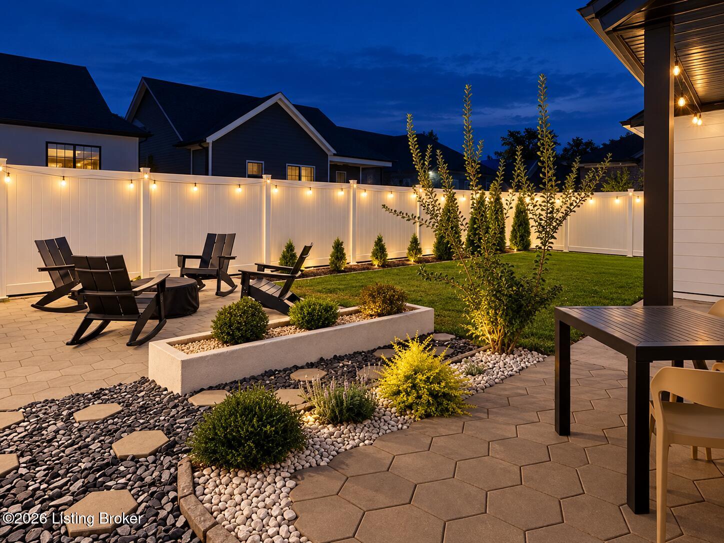 Backyard patio with lounge chairs, a landscaped lawn, and string-lit white fencing at dusk.