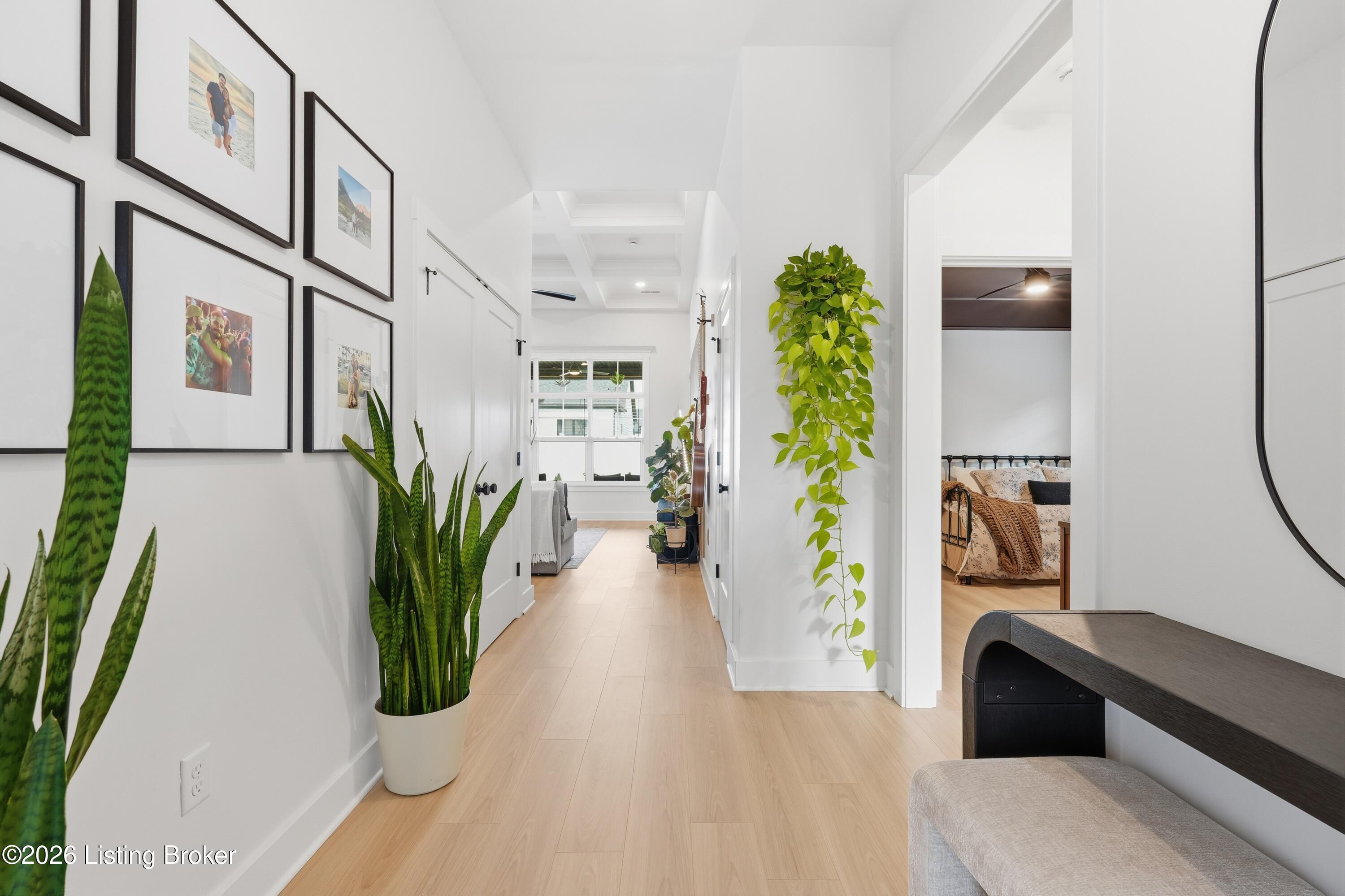 Bright hallway with pale wood flooring, framed photos, potted plants, and an open view into a bedroom.
