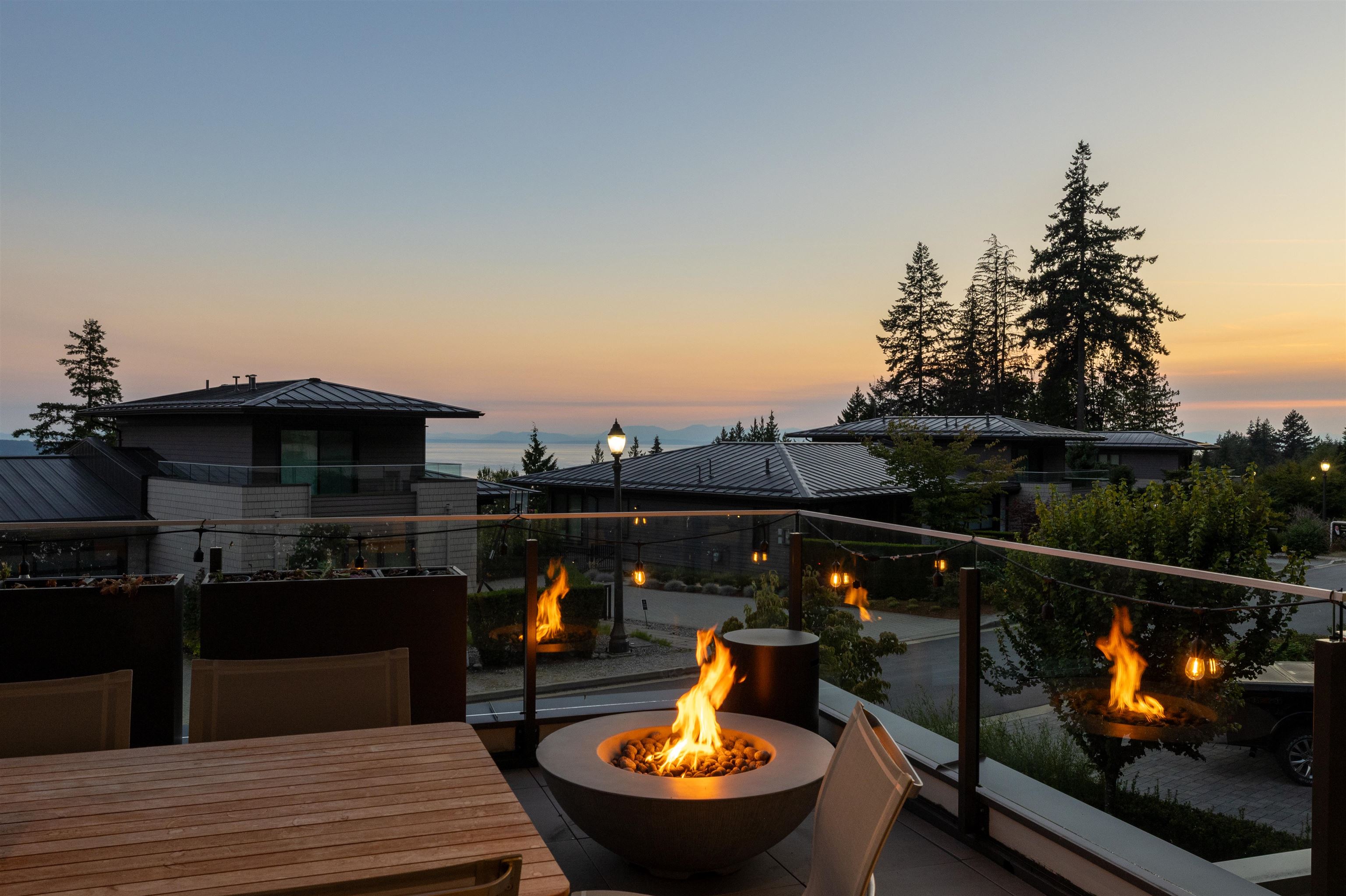 Outdoor terrace with a round fire pit and glass railing overlooking modern homes and distant water.