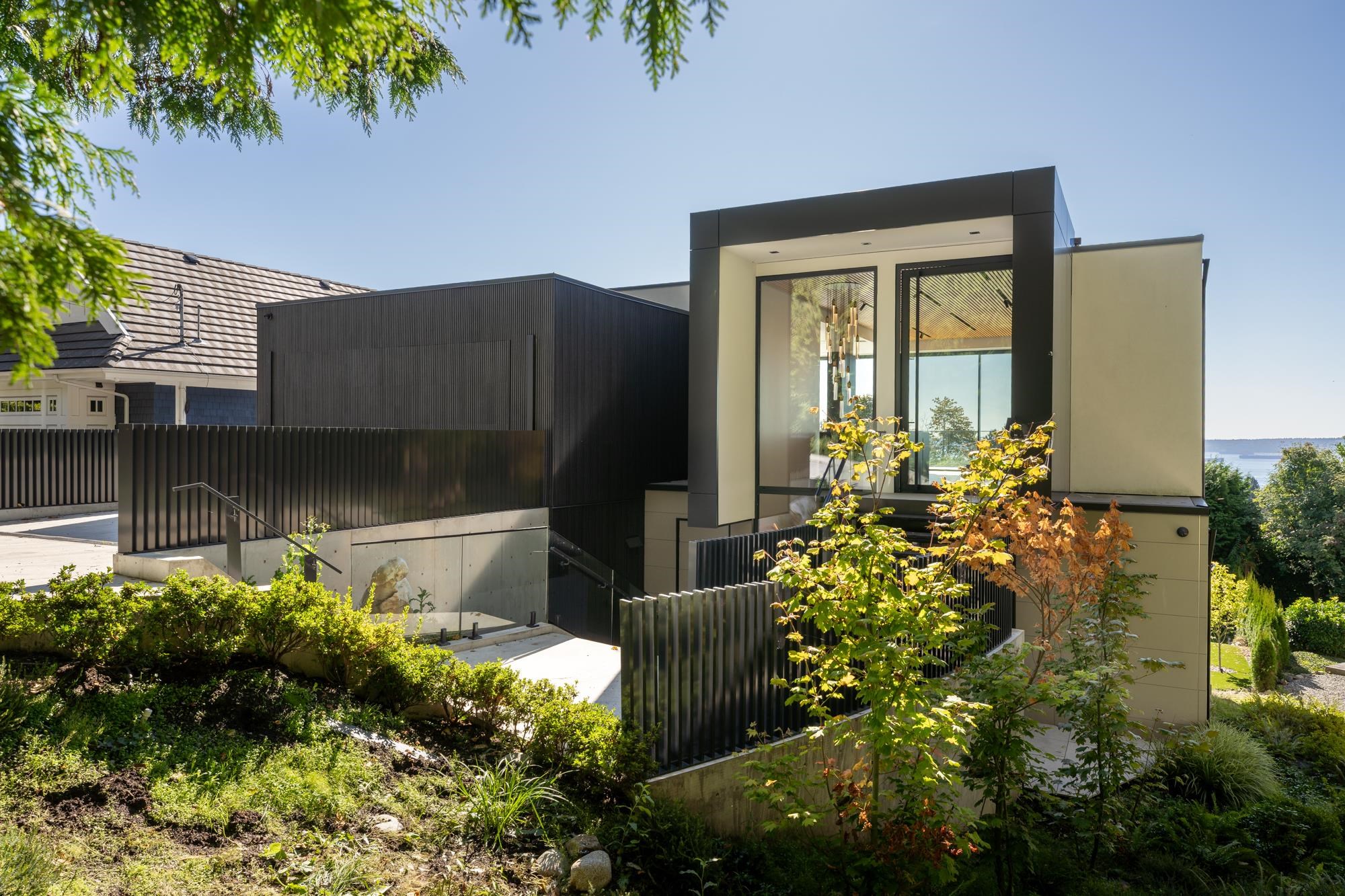 Modern home with dark cladding, floor-to-ceiling windows, landscaped gardens, and a glimpse of water.
