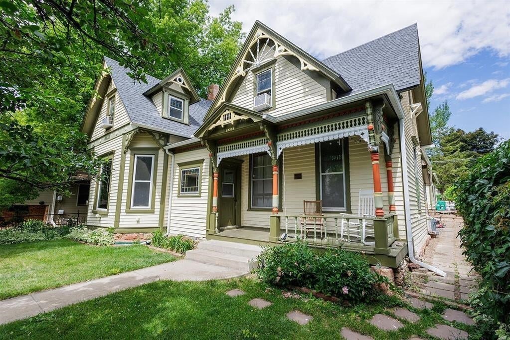 Sage Victorian-style house with ornate porch trim, red columns, and a leafy lawn.