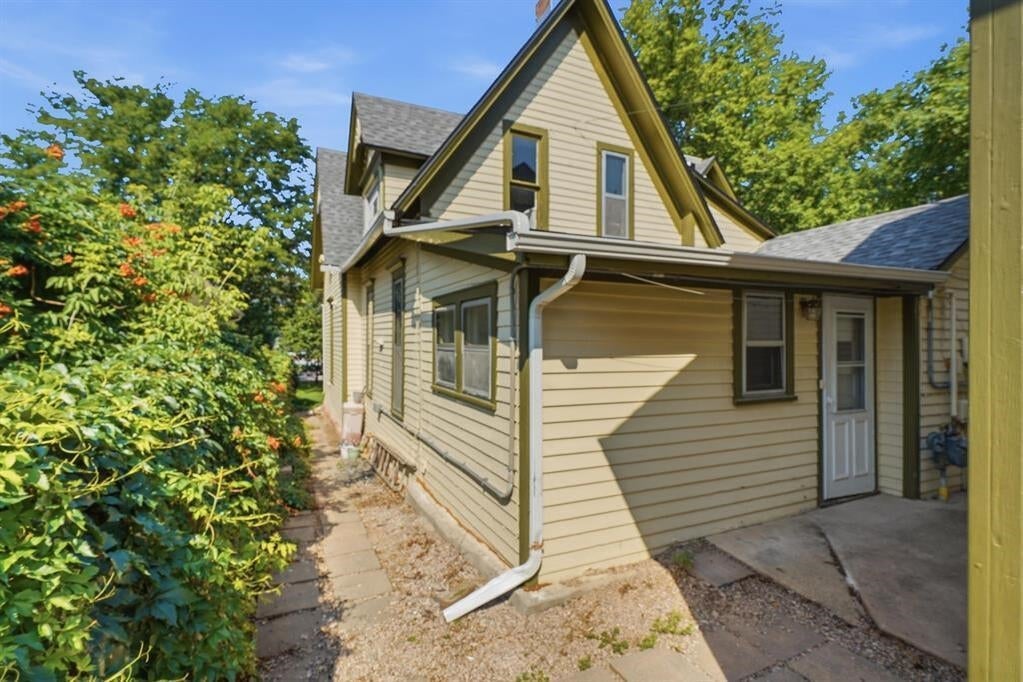 Beige two-story house with a steep gable, side entrance, and leafy shrubs along a paved walkway.