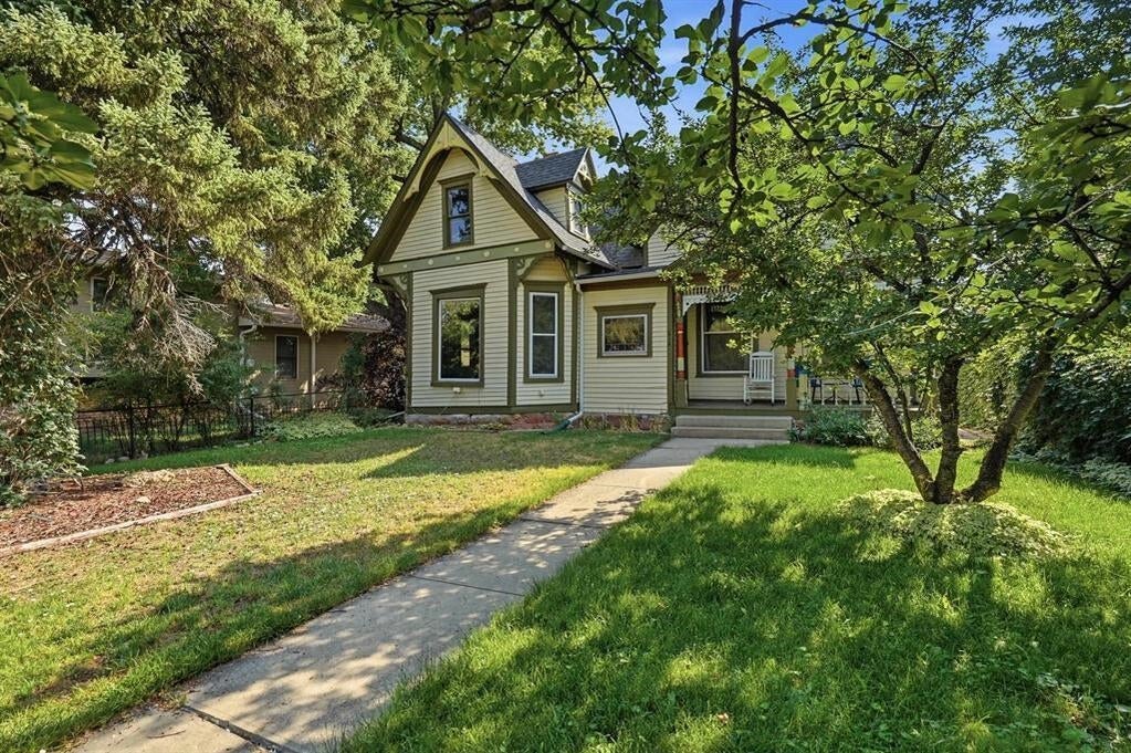 Sage-green gabled house with a covered porch, mature trees, and a walkway through the front lawn.