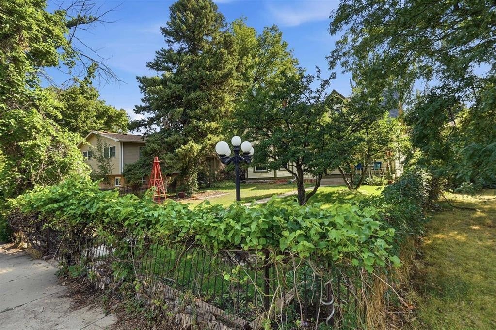 House behind a leafy hedge and decorative fence, with mature trees and a multi-globe yard lamp.