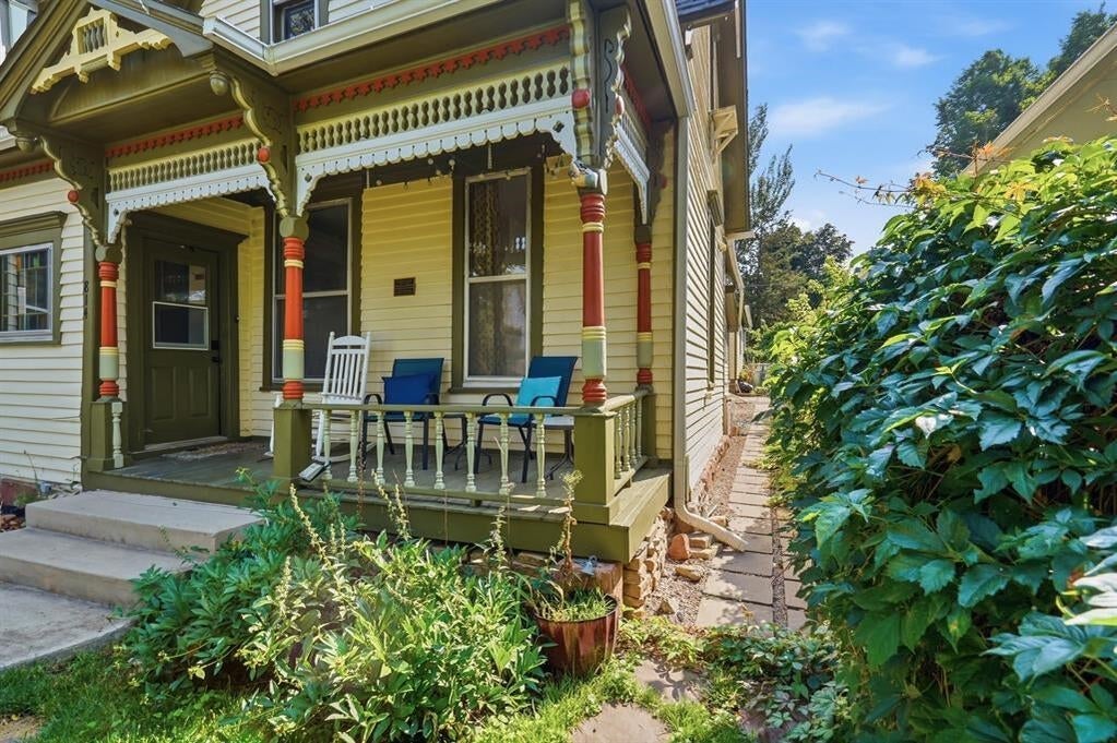 Yellow Victorian-style house with an ornate covered porch, chairs, and a stone path beside lush greenery.
