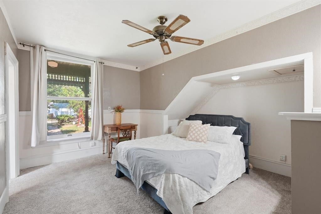 Furnished bedroom with a white bed, dark headboard, sloped ceiling, and large curtained window.