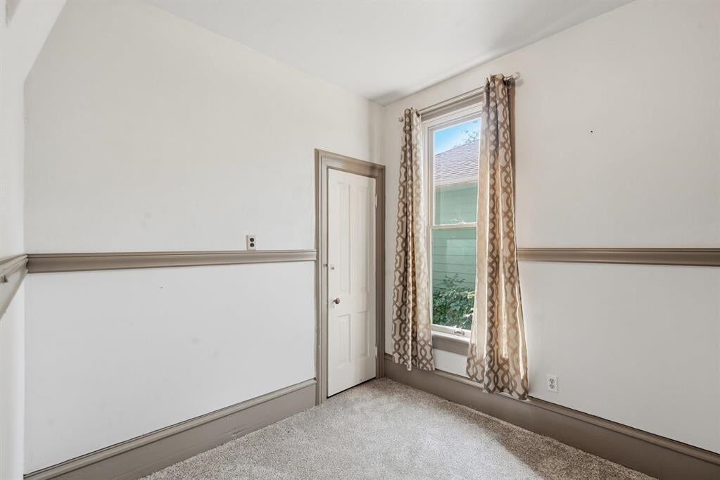 Empty carpeted room with a tall curtained window and paneled door.