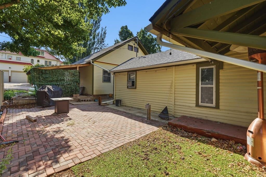 Brick patio beside a yellow-sided home, shaded by mature trees and bordered by a small lawn.