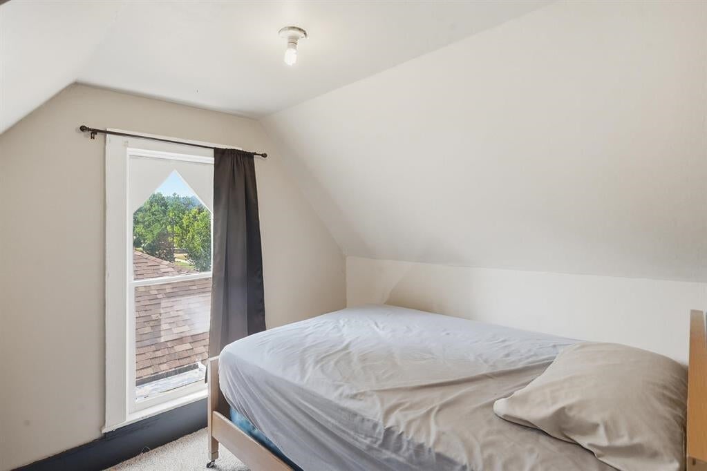 Upstairs bedroom with a sloped ceiling, white-bedded bed, and tall window overlooking trees.