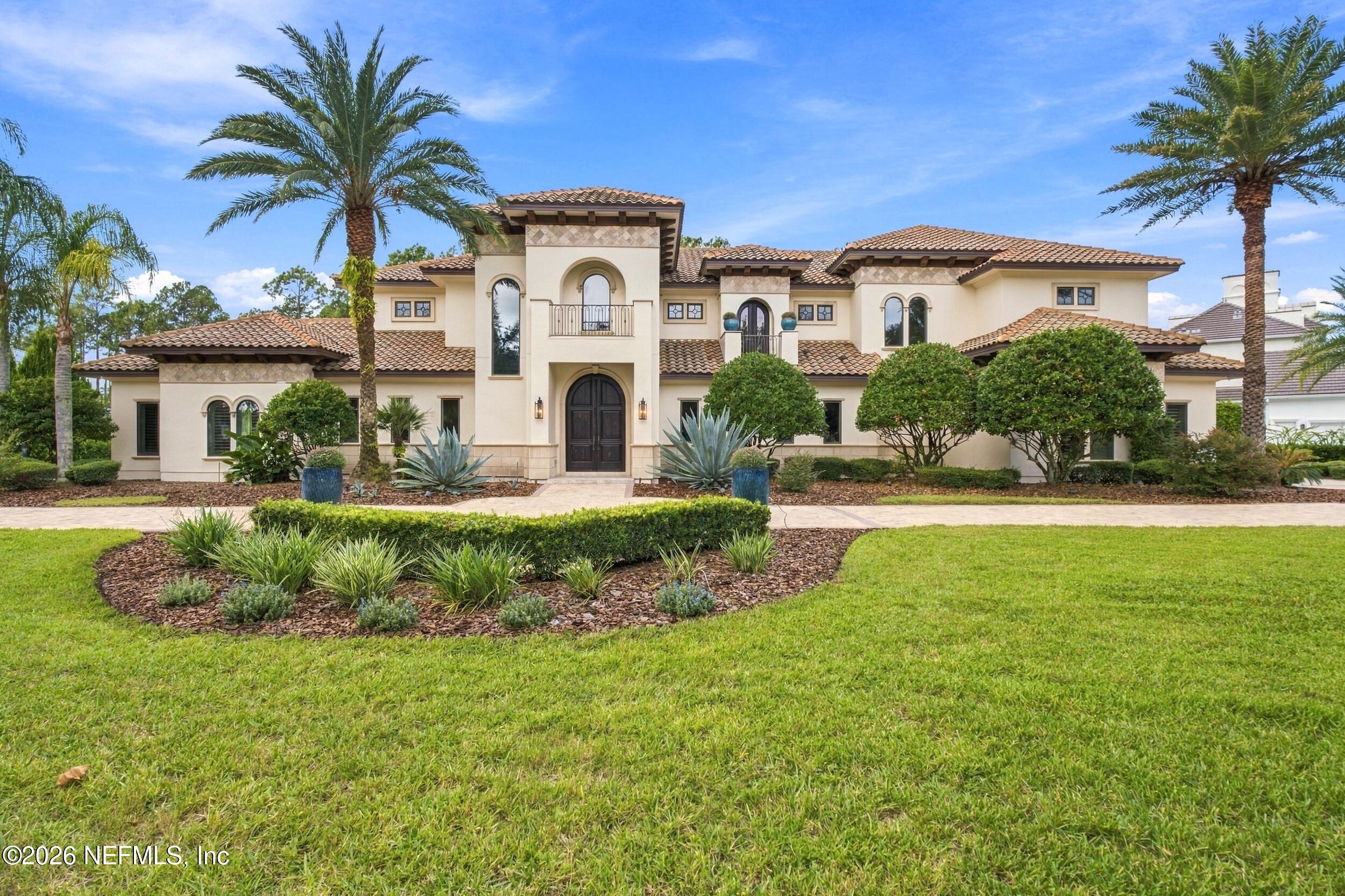 Two-story cream stucco home with a tiled roof, arched entry, palms, and landscaped lawn.