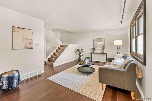 Staged living room with hardwood floors, a sofa, and a staircase beside large windows.