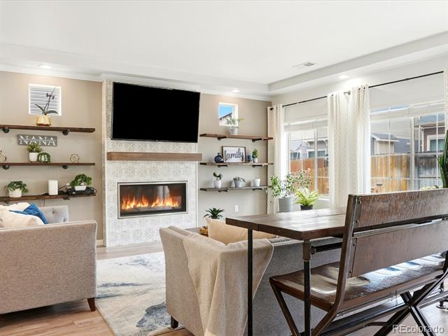 Living and dining area with a tiled fireplace, wall-mounted TV, and large windows overlooking a fenced yard.