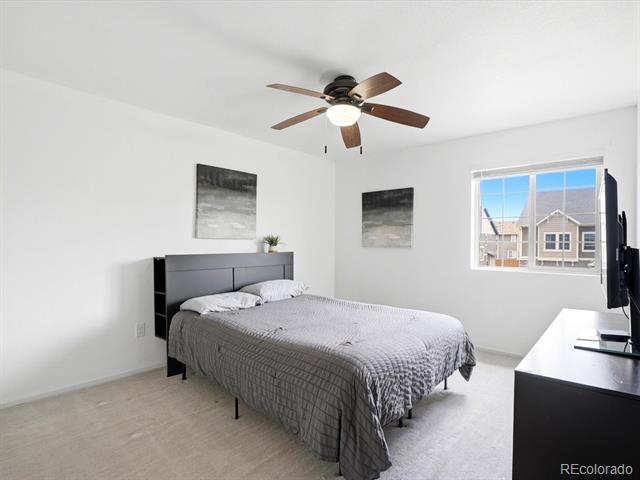 Bedroom with gray bedding, a dark ceiling fan, abstract wall art, and a window overlooking neighboring homes.