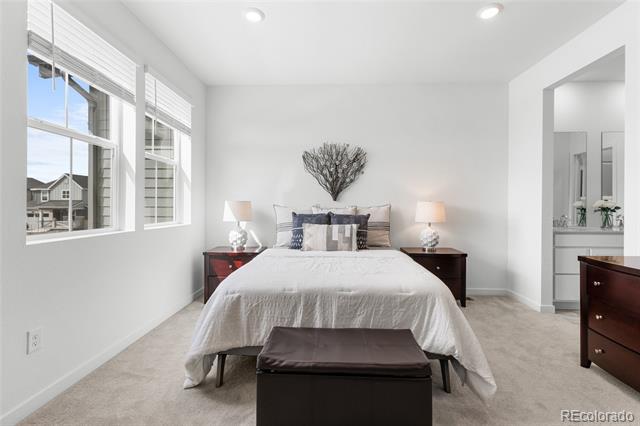 Staged bedroom with a white bed, dark wood nightstands, a cushioned bench, and large windows.