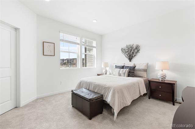 Staged bedroom with a neutral bed, dark wood nightstand and bench, and windows with white blinds.