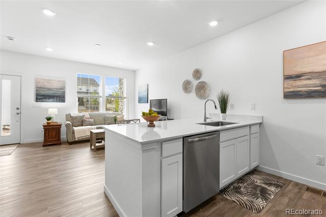 Bright kitchen with a large white island, stainless-steel dishwasher, and adjoining living area.