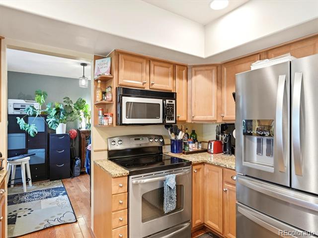 Kitchen with wood cabinets, granite counters, and stainless appliances, opening to a room with a desk and plants.