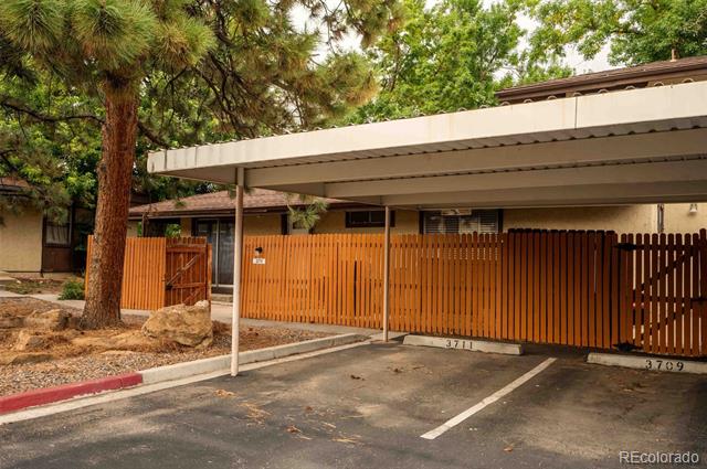 Residence behind a tall wooden fence, with covered parking and a large pine tree beside the driveway.