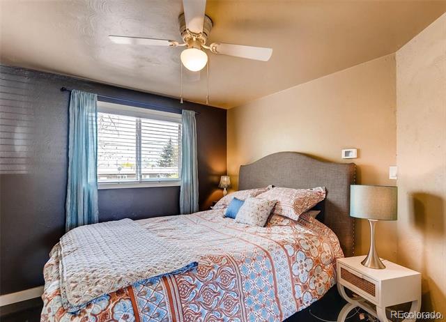 Bedroom with a patterned bedspread, gray upholstered headboard, blue curtains, and ceiling fan.