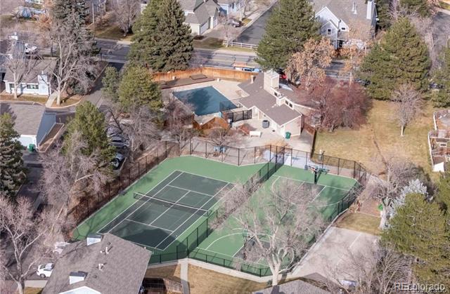 Aerial view of a fenced tennis court and swimming pool among nearby homes.