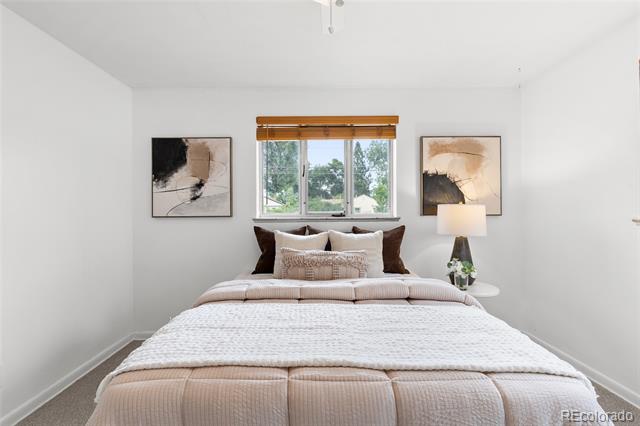 Staged bedroom with a large pale quilted bed, abstract artwork, and a window with wooden blinds.