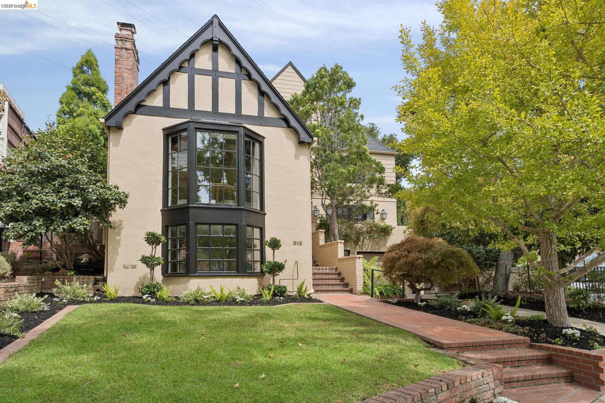 Cream Tudor-style house with dark trim, a tall bay window, and brick paths through the landscaped front yard.