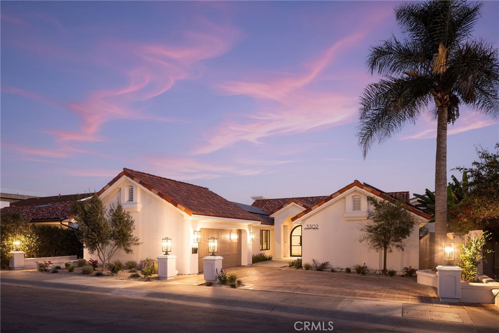 White stucco home with a tiled roof and tall palm tree, warmly lit at dusk.