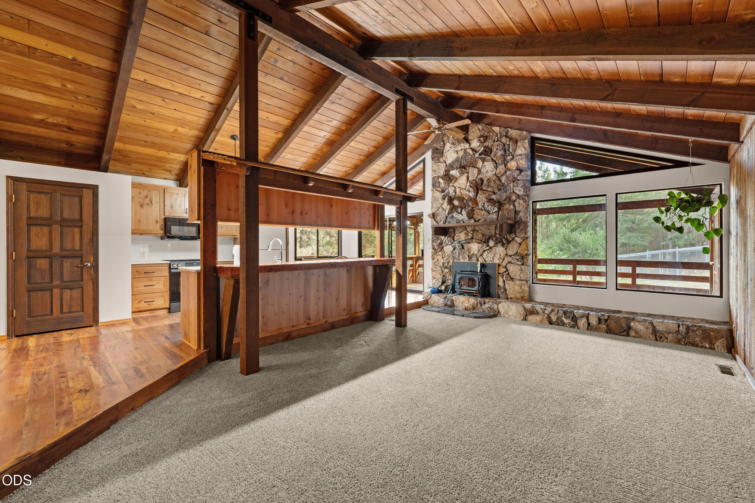 Living room with vaulted wood ceiling, stone fireplace, large windows, and adjoining kitchen.