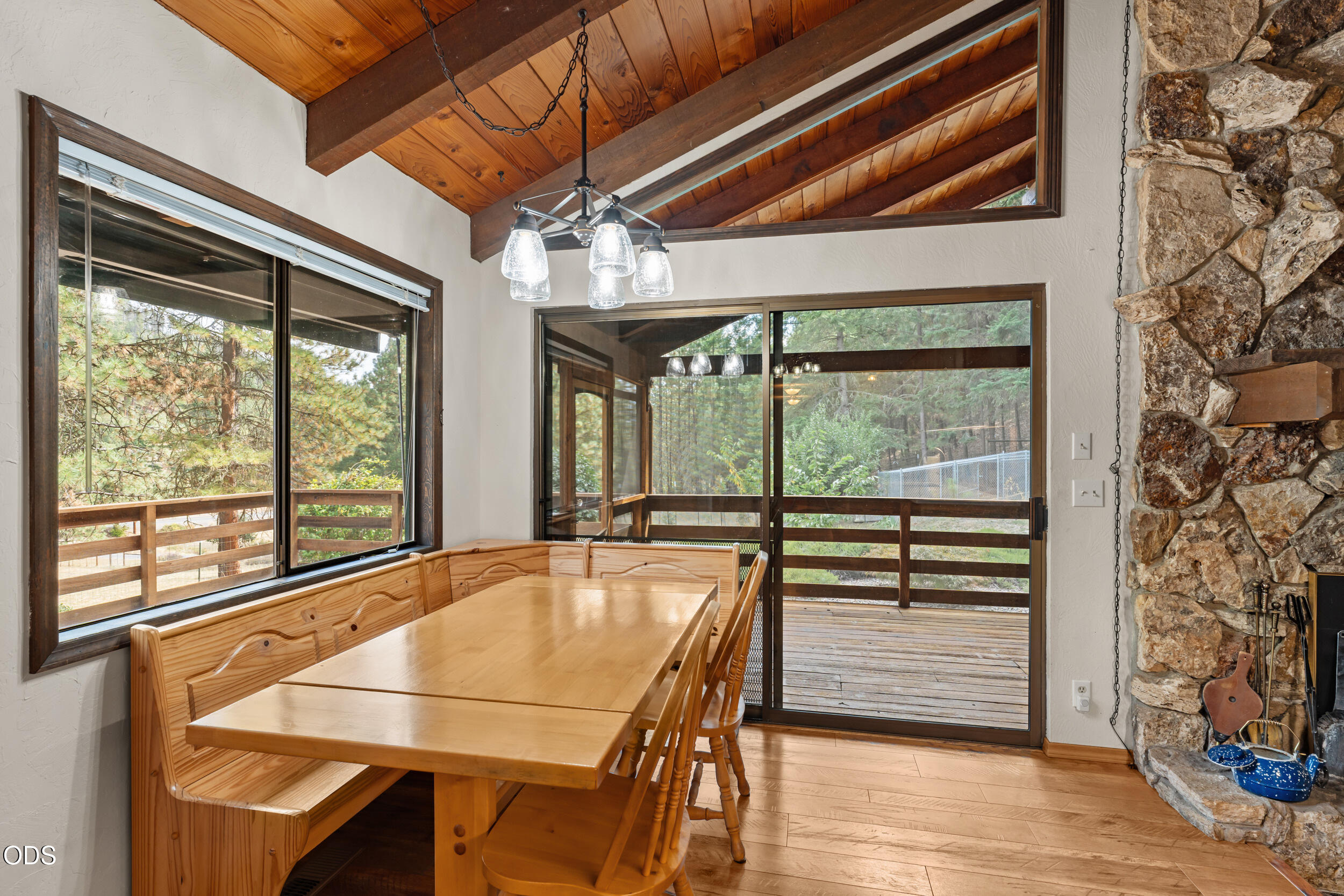 Dining area with a wooden table, vaulted timber ceiling, stone fireplace, and windows overlooking a deck.