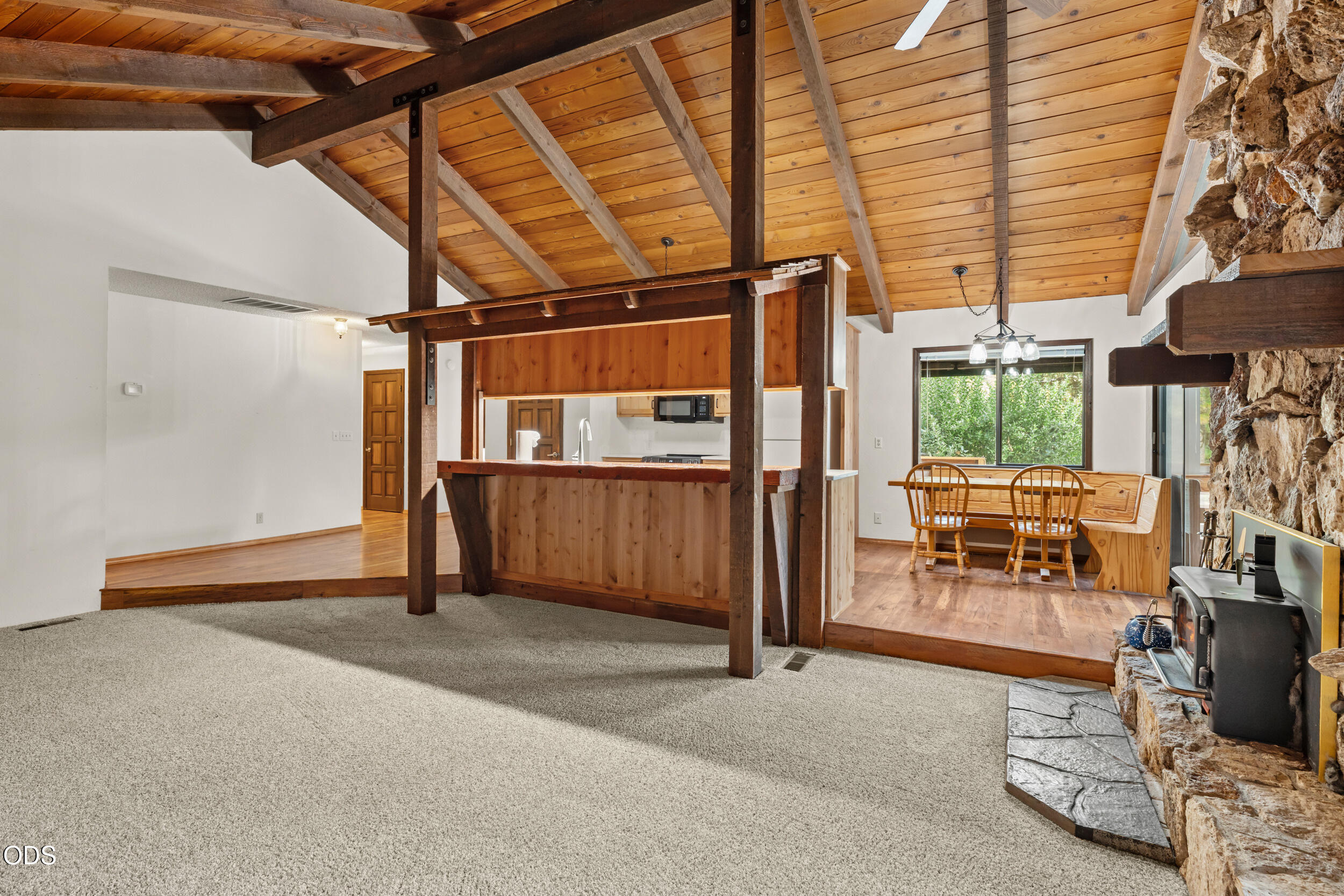 Living room with vaulted wood ceiling, exposed beams, and a stone fireplace beside the dining nook.